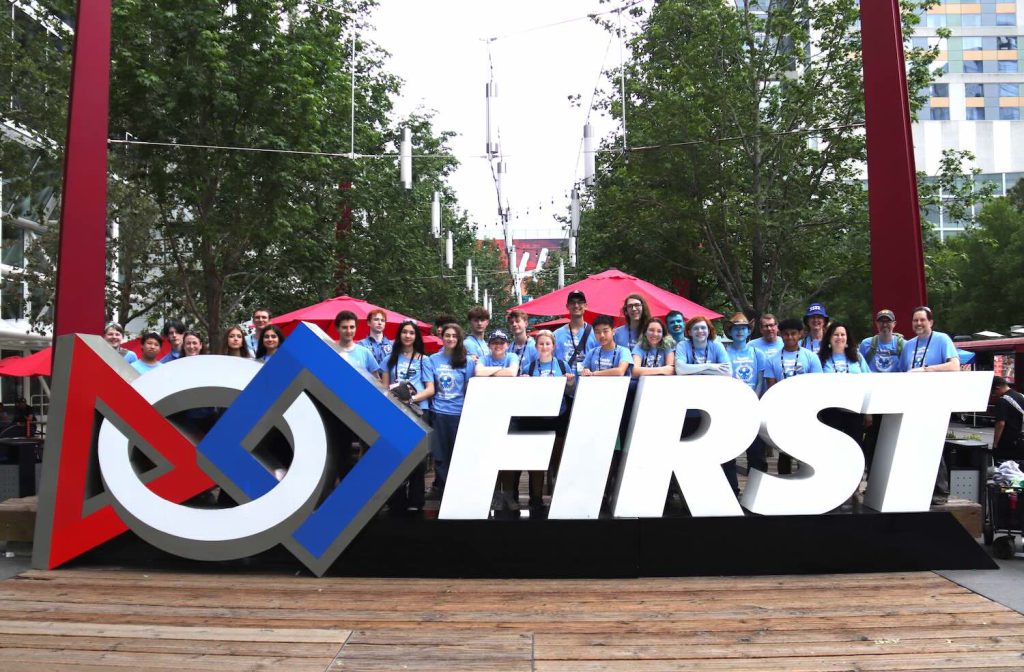Large sculpture sign that says FIRST and has the logo. Team 3322 poses behind it.